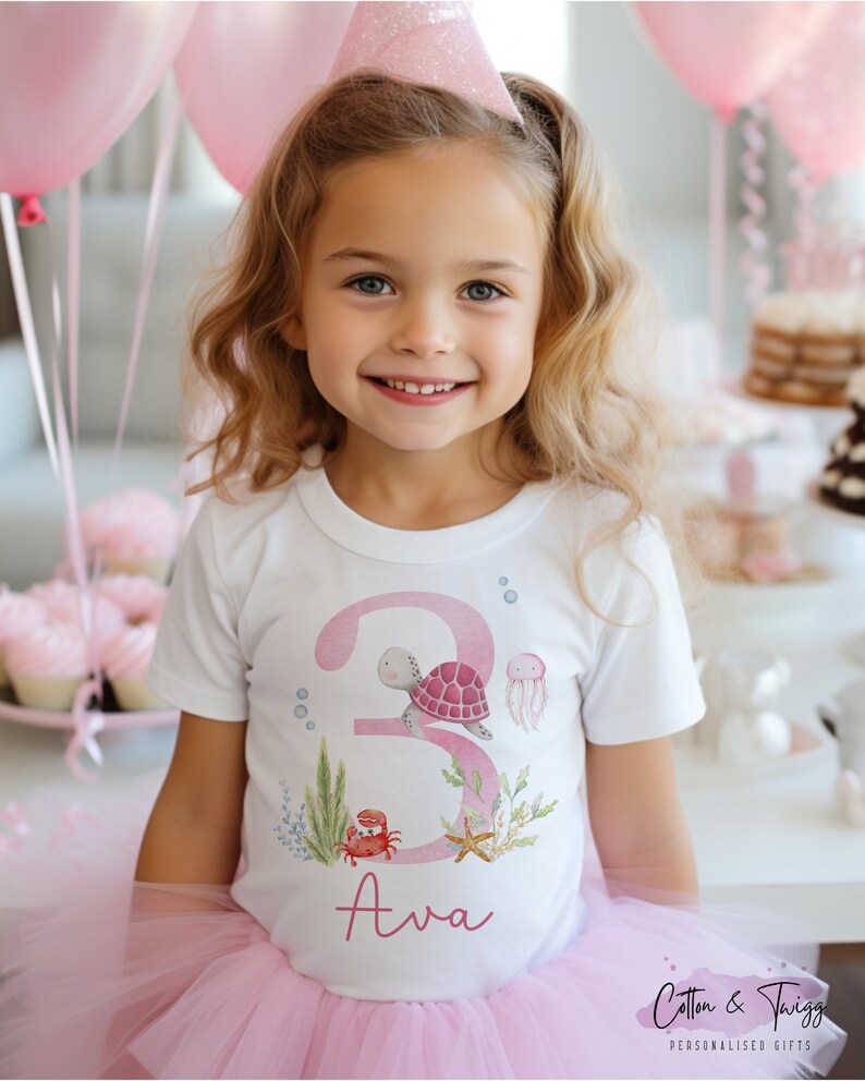 K&ouml;nnte beinhalten: Ein wei&szlig;es T-Shirt mit einer pinkfarbenen Zahl 3, einer Schildkr&ouml;te, einem Krebs, einem Seestern und Algen. Der Name Ava ist in Pink unter der Zahl geschrieben.
