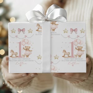 Personalised pink first birthday wrapping paper featuring a cute teddy bear in a tutu holding balloons, rocking horse, gold stars, pink bows, and the number one. Text reads “Happy First Birthday to a Very Special Daughter/Granddaughter/Any relation ” Ideal for baby girl’s first birthday gifts.
