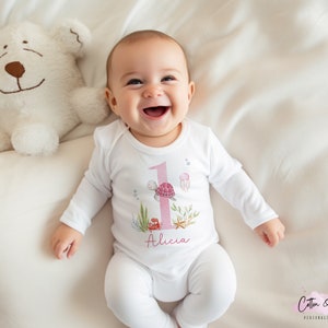 K&ouml;nnte beinhalten: Ein wei&szlig;er Baby-Strampler mit einer pinkfarbenen Nummer eins, einer Schildkr&ouml;te und einem Seestern-Design. Der Strampler tr&auml;gt den Namen "Alicia".
