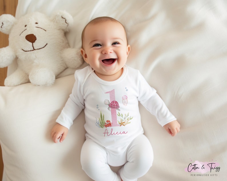 K&ouml;nnte beinhalten: Ein wei&szlig;er Baby-Strampler mit einer pinkfarbenen Nummer eins, einer Schildkr&ouml;te und einem Seestern-Design. Der Strampler tr&auml;gt den Namen "Alicia".