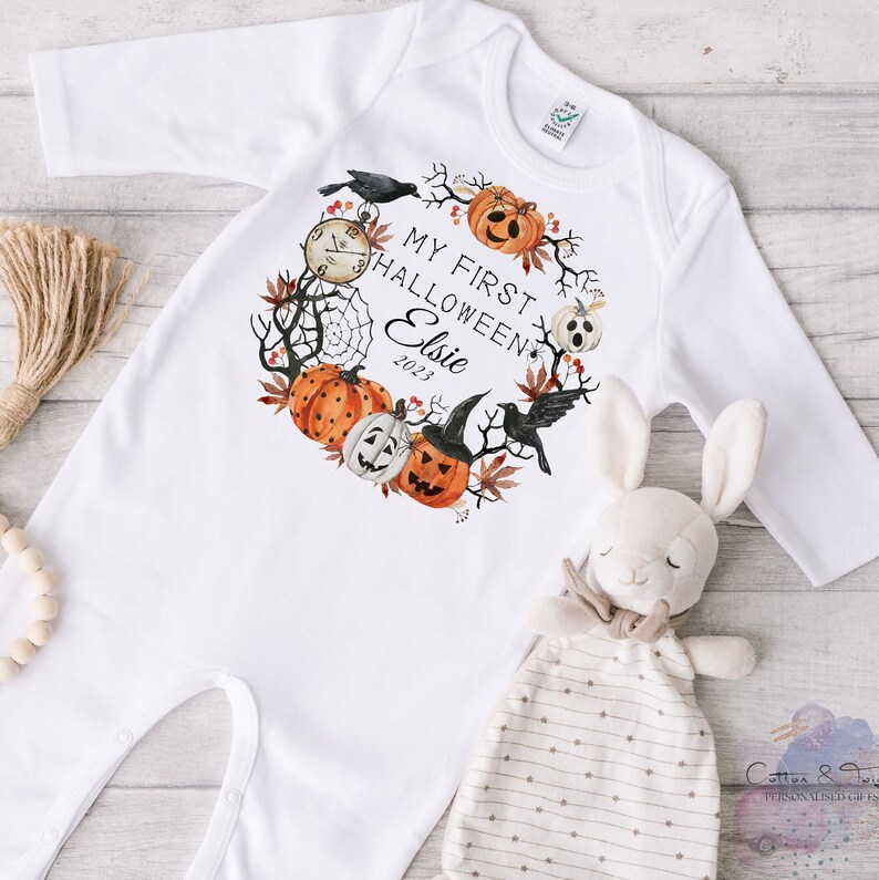 May include: White baby onesie with long sleeves, featuring a Halloween-themed wreath design. The wreath includes pumpkins, a clock, a spiderweb, and the text "My First Halloween Elsie 2023". A stuffed bunny and a bundle of twine are also in the image.