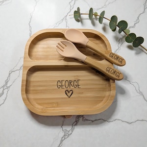 May include: A bamboo baby plate with two compartments and a matching spoon and fork set. The plate and utensils are engraved with the name "GEORGE" and a heart symbol. The spoon and fork have a light pink silicone tip.