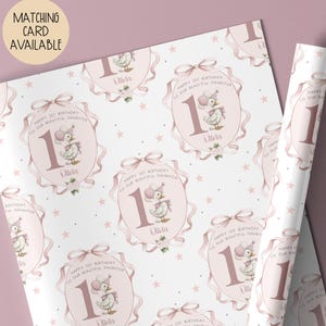 May include: White wrapping paper with a repeating pattern of pink oval frames, featuring a duck wearing a party hat and the text "Happy 1st Birthday to our beautiful daughter Olivia". Pink bows and stars complete the design.
