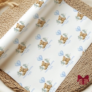 Puede incluir: Papel de regalo blanco con un patrón repetido de un oso de peluche marrón que sostiene globos azules en forma de corazón. El texto "Happy 1st Birthday Teddy" se repite en todo el patrón.