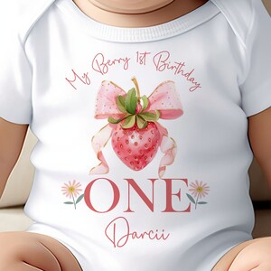 May include: White baby bodysuit with a strawberry design and the text "My Berry 1st Birthday". The design includes a pink bow, a strawberry, and the word "ONE". The name "Darcii" is also printed on the bodysuit.