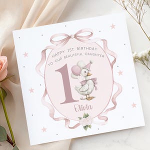 May include: A square greeting card with a pink and white design. The card features a watercolor illustration of a duck holding balloons, a large number 1, and the text "Happy 1st Birthday to our beautiful daughter Olivia."