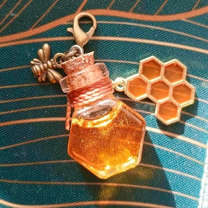 Può includere: Un ciondolo con una piccola bottiglia esagonale di vetro riempita di liquido ambrato, probabilmente miele. La bottiglia ha un tappo di sughero ed è avvolta con filo color rame. Un ciondolo a forma di ape e favo color oro è attaccato.