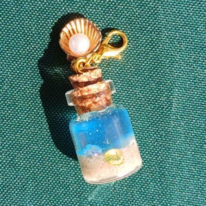 May include: A miniature glass bottle charm filled with blue liquid and sand, topped with a cork and a gold-colored seashell with a pearl. The charm is attached to a gold-colored clasp.
