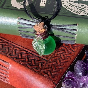 May include: A green glass bottle pendant with a cork stopper and a green leaf charm. The bottle is hanging from a black cord necklace. The necklace is resting on a brown leather book with a Celtic knot design.