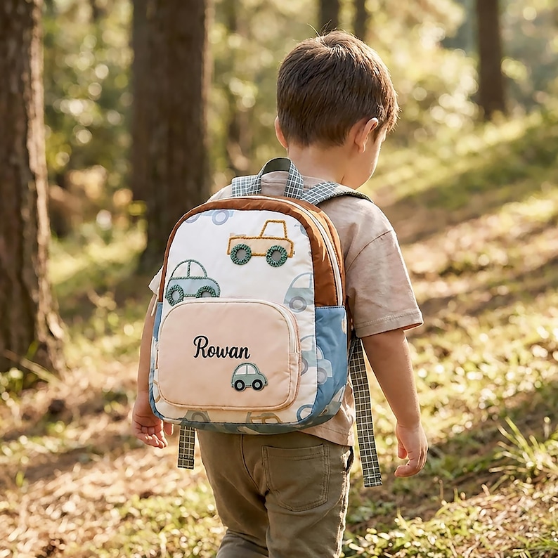 May include: A small backpack featuring a white fabric body with a repeating pattern of embroidered cars in shades of blue and tan. The front pocket is solid light tan with the name Rowan embroidered in black script and a small car icon.