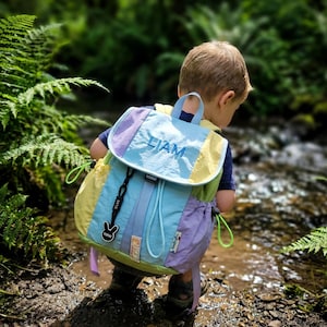 May include: A multi-coloured backpack with the name "LIAM" embroidered on the flap. The backpack has blue, yellow, and purple panels, a black buckle, and a drawstring closure. The backpack is being worn by a child.