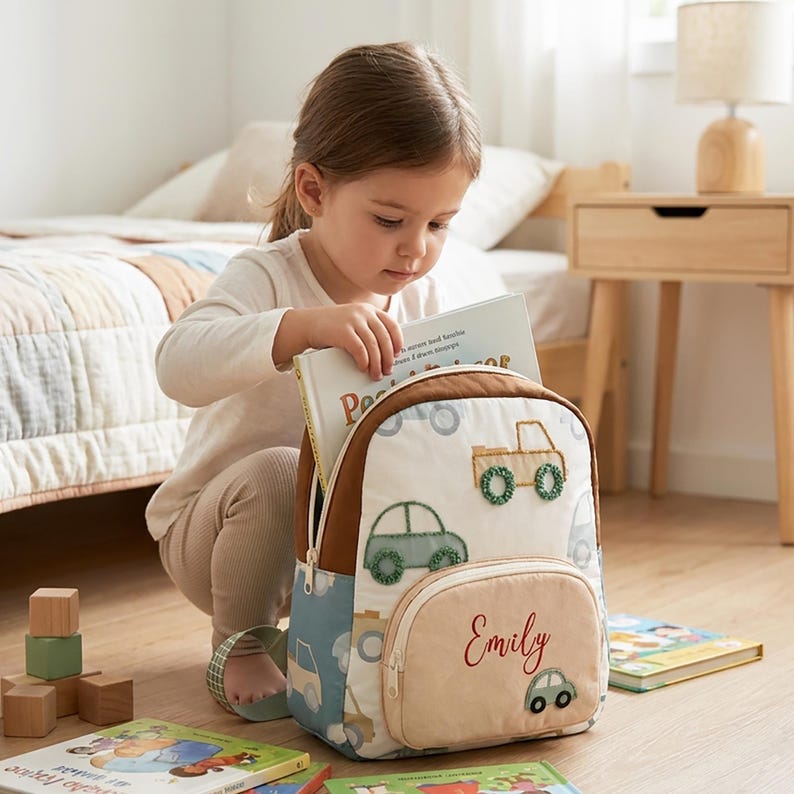 May include: A white backpack featuring embroidered green and tan car motifs and a tan front pocket with the name Emily stitched in red script. The backpack has brown trim, a light blue side panel with car prints, and a white zipper closure.