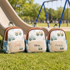 Può includere: Tre zaini personalizzati con disegni di auto. Ogni zaino ha una parte superiore marrone, un corpo bianco con illustrazioni di auto e una tasca beige con un nome. I nomi sono "Emily", "Olivia" e "Grace". Gli zaini sono sull'erba verde.
