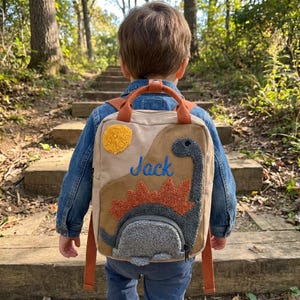 May include: A tan and brown backpack with a dinosaur design and the name "Jack" embroidered in blue. The backpack features orange straps and a yellow sun applique. The child is wearing a denim jacket.