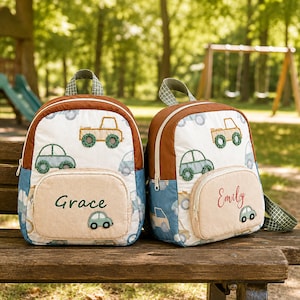 May include: Two quilted backpacks featuring a white base with a pattern of green and tan cars. Each bag has a tan front pocket with a name embroidered in green or red, and a small car appliqu&eacute;. The backpacks have brown trim and green handles.