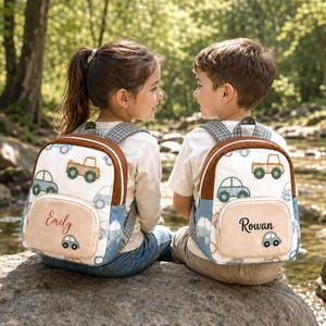 May include: Two white backpacks featuring a repeating pattern of light blue and green cars and trucks. Each backpack has a brown trim, a front pocket with a personalized name embroidered in black or red script, and grey plaid shoulder straps.
