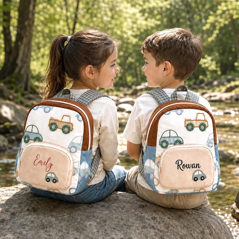 May include: Two white backpacks featuring a repeating pattern of light blue and green cars and trucks. Each backpack has a brown trim, a front pocket with a personalized name embroidered in black or red script, and grey plaid shoulder straps.