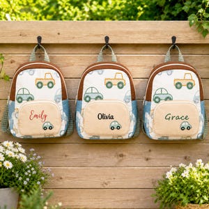 May include: Three backpacks with a white base featuring a repeating pattern of green and yellow cars. Each bag has a tan front pocket personalized with the names Emily, Olivia, and Grace, and a small embroidered car icon on the pocket.