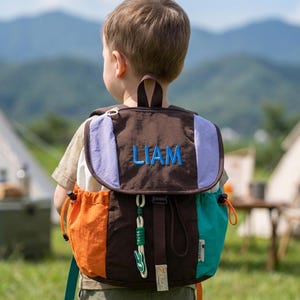 May include: A multi-coloured backpack with the name "LIAM" embroidered in blue. The backpack has a brown flap with a light purple accent, an orange and teal front pocket, and a drawstring closure. It has a top handle and adjustable straps.