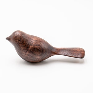 May include: A dark brown wooden bird figurine with a smooth, polished finish. The bird is carved in a simple, realistic style with a rounded body and a long, slender tail.