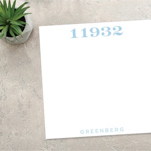 May include: A white notepad with a blue number "11932" and the word "GREENBERG" printed on it. The notepad is on a light gray textured surface.