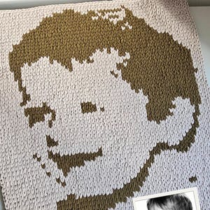 Op de afbeelding: Een vierkante gebreide deken met een gepixeleerd, tweekleurig portret van een jong kind. Het ontwerp gebruikt olijfgroen garen tegen een lichtcrème achtergrond om de gelaatstrekken en het haar van het kind op de kleine foto na te bootsen.