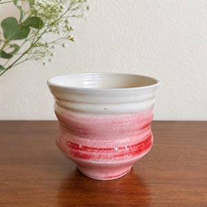 May include: A ceramic cup featuring a wide, flared rim and a rounded body. The upper portion is finished in a smooth off-white glaze, while the lower section transitions into a vibrant, textured pink and red gradient with visible horizontal ridges.