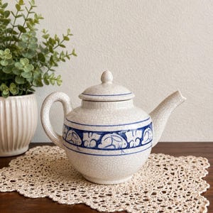 Può includere: Una teiera in ceramica con finitura smaltata color crema effetto crepato, caratterizzata da una fascia orizzontale con illustrazioni di conigli dipinte a mano in blu. La teiera include un coperchio abbinato con pomello.