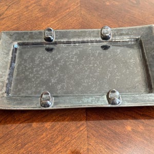 May include: A rectangular tray with a dark, mottled metallic finish. The tray features a wide, flat rim and four small, raised, rounded metallic accents positioned at the corners of the inner rectangular base, creating a distinct, textured appearance.