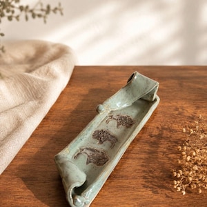 May include: A rectangular ceramic tray with a light green glaze and rolled edges. The interior surface features three embossed brown bison figures arranged in a line along the center of the elongated dish, which has a smooth, glossy finish.