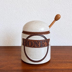 Può includere: Un vasetto per miele cilindrico in ceramica con smalto bianco sporco maculato e bordo marrone scuro. La parte anteriore presenta un emblema circolare marrone in rilievo con la parola HONEY in lettere maiuscole. Include coperchio e mestolo in legno.