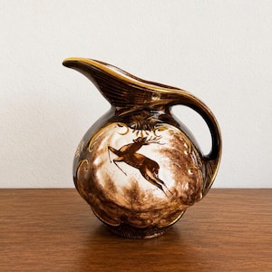 May include: A dark brown ceramic pitcher featuring a glossy finish and a gold-trimmed rim and handle. The central design displays a leaping deer in a lighter brown tone, surrounded by ornate, swirling gold-colored decorative accents.
