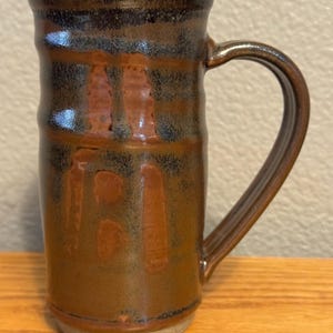 Può includere: Una tazza in ceramica fatta a mano con smalto marrone e nero. La tazza ha una forma cilindrica con un manico curvo. L'esterno presenta una superficie strutturata con variazioni di colore, creando un aspetto rustico. La base è non smaltata, rivelando il colore naturale dell'argilla.