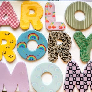 Puede incluir: Una colección de once letras y formas decorativas dispuestas para deletrear ARLO RORY MOM. Cada pieza presenta un patrón único que incluye cuadros rosas, amarillo sólido, cerezas rojas, rayas verdes, caras sonrientes amarillas, arcoíris, estampado de leopardo, hojas de palma, lavanda sólido, arcos azul claro y cuadros de flores en blanco y negro.