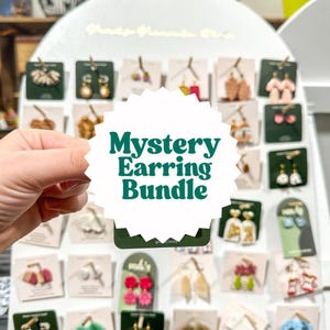 May include: A display of various earrings on small cards, with a white sign that reads "Mystery Earring Bundle" in green text. The earrings are in different styles and colors, including gold, pink, and white.