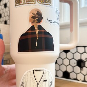 May include: A white stainless steel tumbler with a pink handle. The tumbler has several stickers on it, including a glitter sticker that says "chiefs", a sticker of a woman with a braid, a sticker of a butterfly, and a sticker of a white sweater with a black collar and buttons. The sticker with the woman also has the text "long story short" on it.