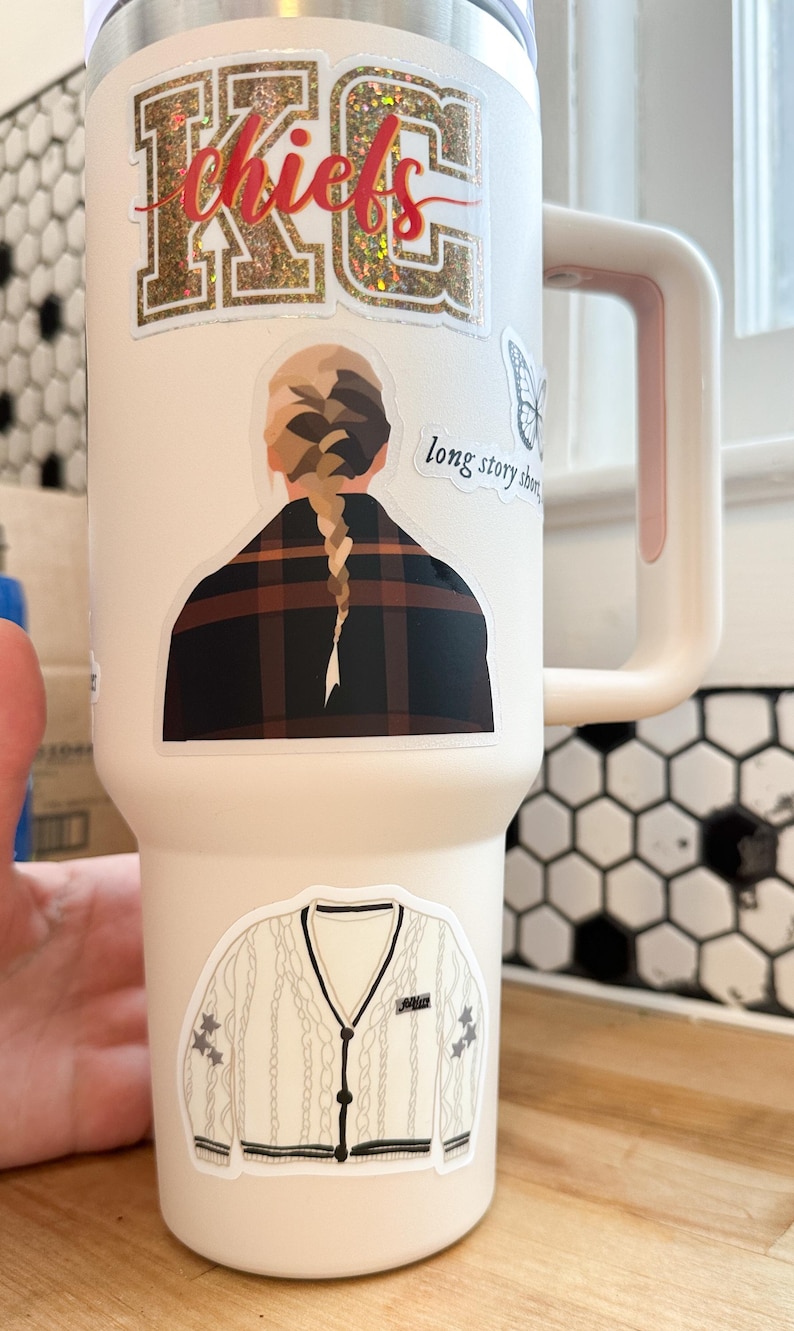 May include: A white stainless steel tumbler with a pink handle. The tumbler has several stickers on it, including a glitter sticker that says "chiefs", a sticker of a woman with a braid, a sticker of a butterfly, and a sticker of a white sweater with a black collar and buttons. The sticker with the woman also has the text "long story short" on it.