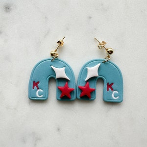 May include: A pair of blue and white earrings shaped like arches with red stars and the letters "KC" on each earring.