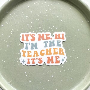 May include: A white sticker with the text "It's me, hi I'm the teacher It's me" in a retro font with orange, yellow, and blue colors.