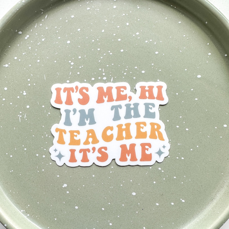 May include: A white sticker with the text "It's me, hi I'm the teacher It's me" in a retro font with orange, yellow, and blue colors.