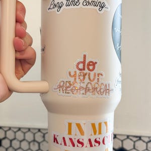 May include: A light pink stainless steel tumbler with a black handle. The tumbler has several stickers on it, including "It's been a long time coming...", "Do your research", and "In my Kansas City Era".