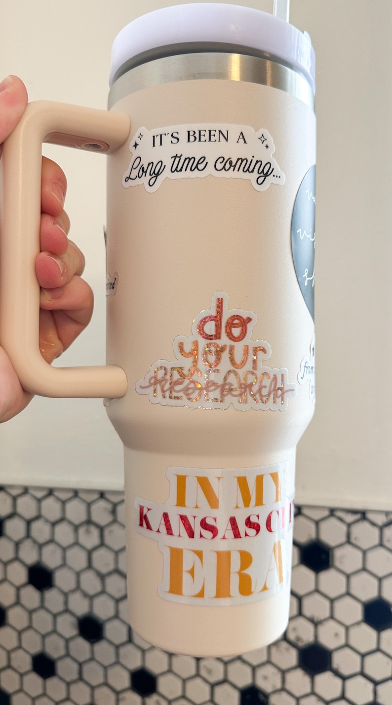 May include: A light pink stainless steel tumbler with a black handle. The tumbler has several stickers on it, including "It's been a long time coming...", "Do your research", and "In my Kansas City Era".