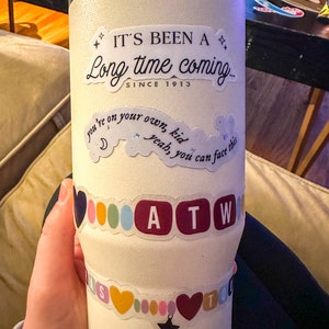 May include: White stainless steel tumbler with various stickers. The stickers include the text "It's been a long time coming... since 1913", "you're on your own, kid, yeah, you can face this", and "ATW".