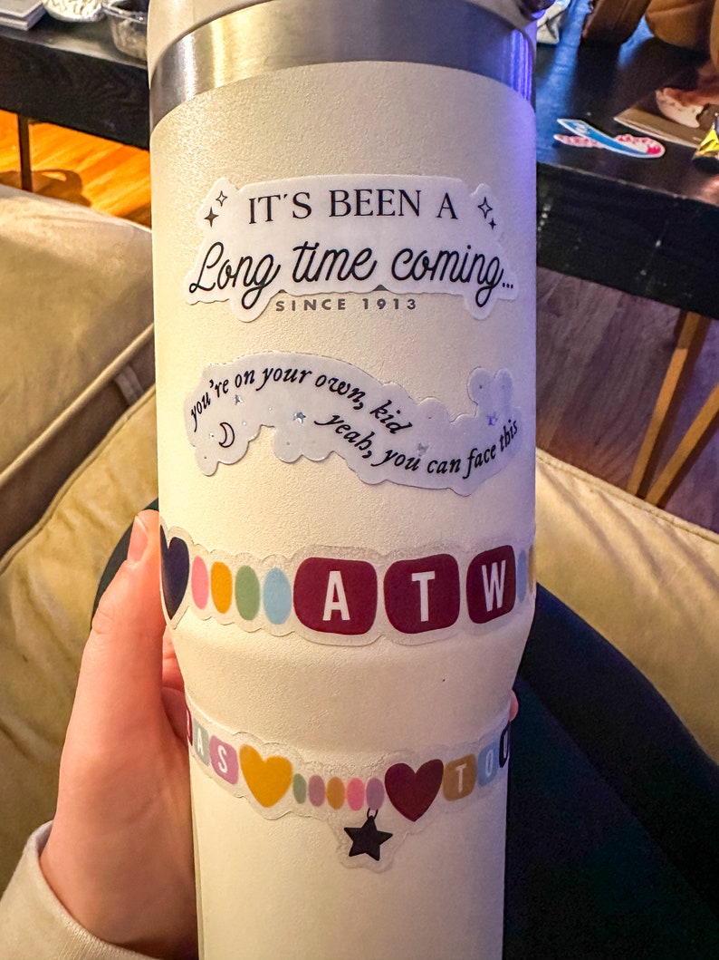May include: White stainless steel tumbler with various stickers. The stickers include the text "It's been a long time coming... since 1913", "you're on your own, kid, yeah, you can face this", and "ATW".