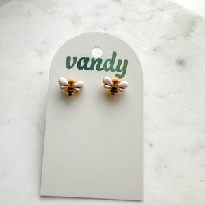 Könnte beinhalten: Ohrstecker in Bienenform auf einer weißen Karte. Die Ohrringe haben goldfarbene Körper, schwarze Streifen und weiße Flügel. Auf der Karte steht das Wort "vandy" in grüner Schrift.