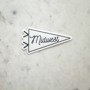 Può includere: Un adesivo bianco a forma di gagliardetto con la scritta "Midwest" in corsivo nero. L'adesivo ha un bordo nero ed elementi decorativi a forma di freccia sul lato sinistro. L'adesivo è su una superficie di marmo bianco.