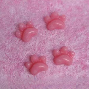 May include: Four small, pink paw-shaped embellishments. The paw prints are a glossy, light pink color and are arranged on a textured, pink surface. These decorative elements are likely used for crafts or embellishments.