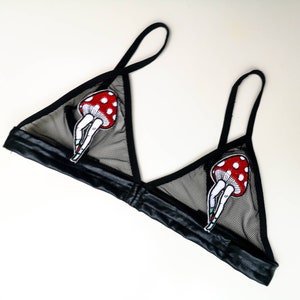 May include: Black mesh bralette with a satin band and straps. The bralette features two mushroom-shaped patches with red caps and white spots, each with legs and high heels. The bra has a triangle shape.