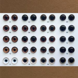 Może przedstawiać: Kolekcja dwudziestu szklanych oczu dla lalek w różnych odcieniach brązu, czerni i przezroczystości. Każde oko jest ponumerowane od 01 do 20. Oczy ułożone są w rzędach i kolumnach na białej powierzchni, z brązowym tłem.