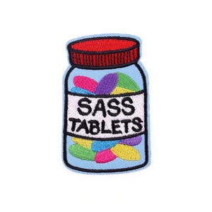 Puede incluir: Un parche azul claro con forma de frasco de pastillas con tapa roja. La etiqueta dice "SASS TABLETS" en texto negro. El frasco está lleno de pastillas oblongas de colores en tonos rosa, morado, amarillo y verde.